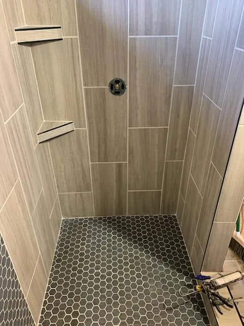 Wood-look tile shower walls with corner shelves and a dark hexagon tile floor